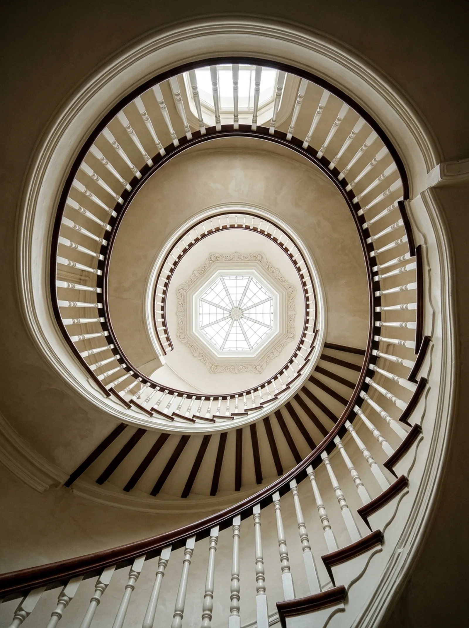 Looking up a restored three-storey Victorian spiral stairwell, white balusters and a mahogany handrail curling towards an octagonal roof lantern